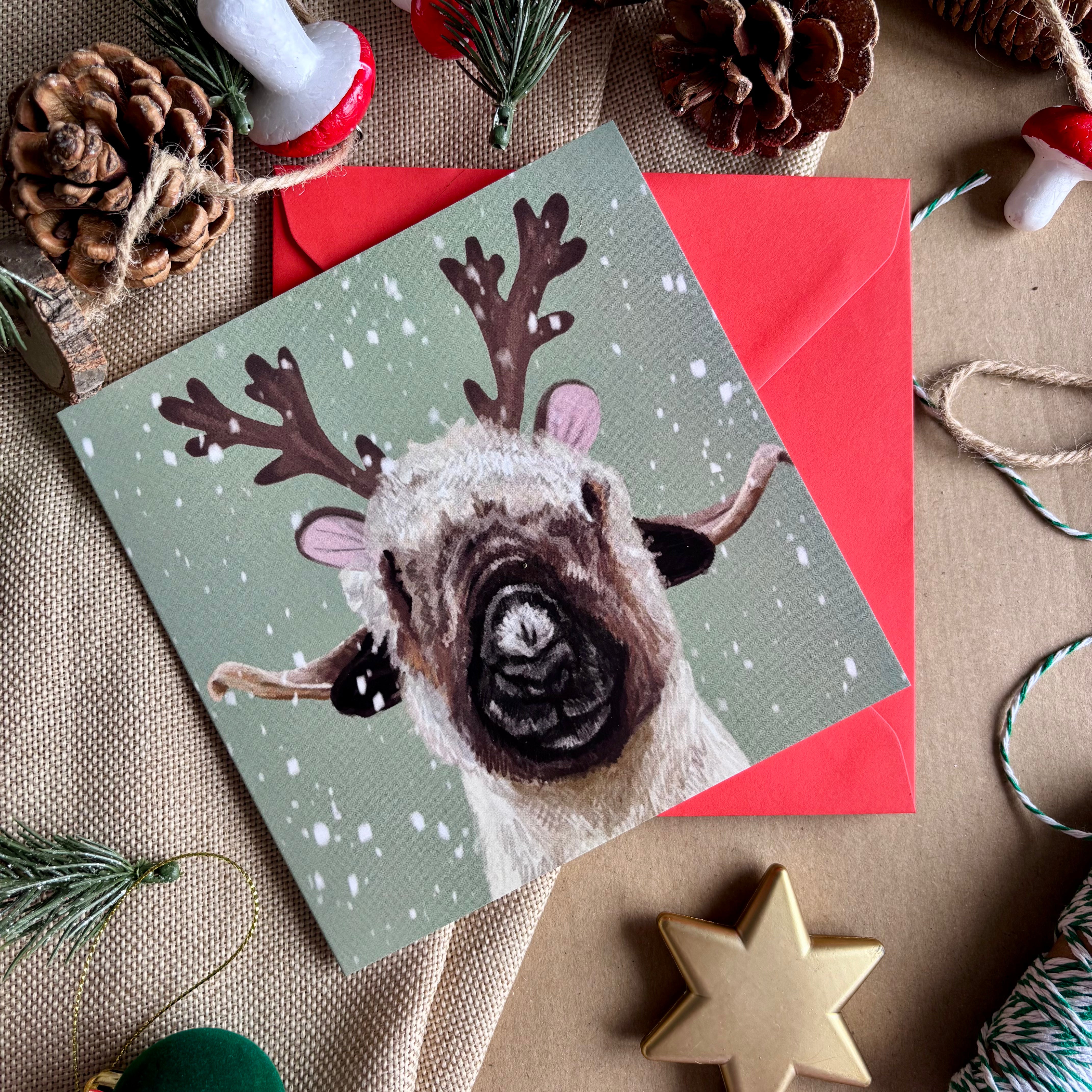 Christmas card with a sheep wearing reindeer antlers on a red envelope, surrounded by festive decorations.